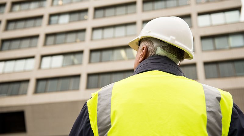 Image of the back of a construction worker with a high visibility vest and hard hat on facing a large building to support Midlands construction article