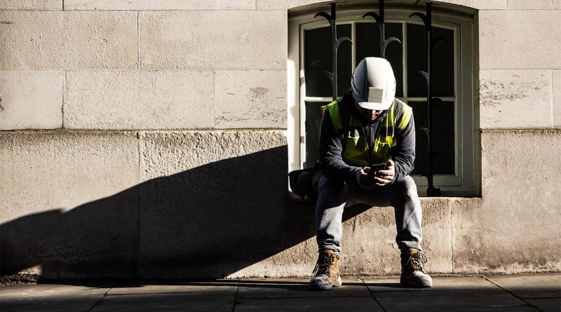 Image of a male construction worker sitting in a window sill on his own looking down at his phone to support UK construction industry article