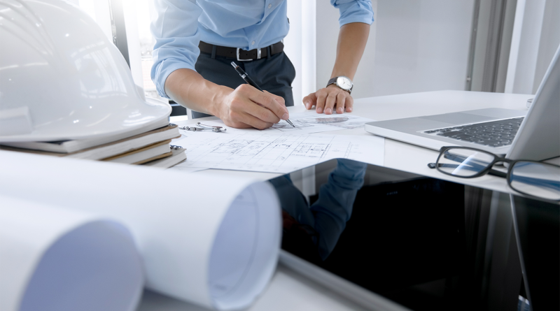 Image of a business professional at a desk with a hard hat, building plans and various different papers on to support construction management article
