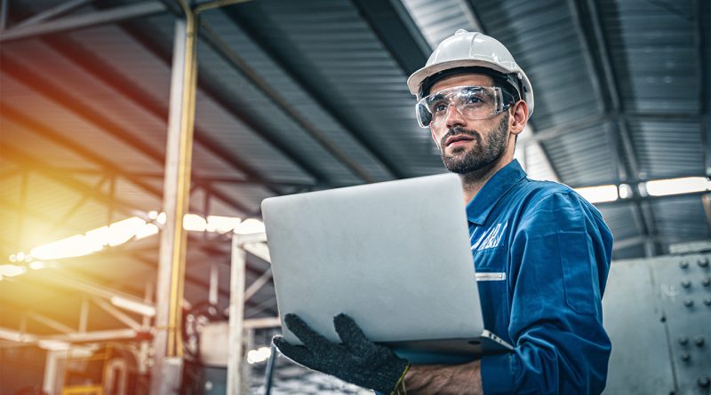 engineer in blue jumpsuit holding laptop in a warehouse to support engineering salary article