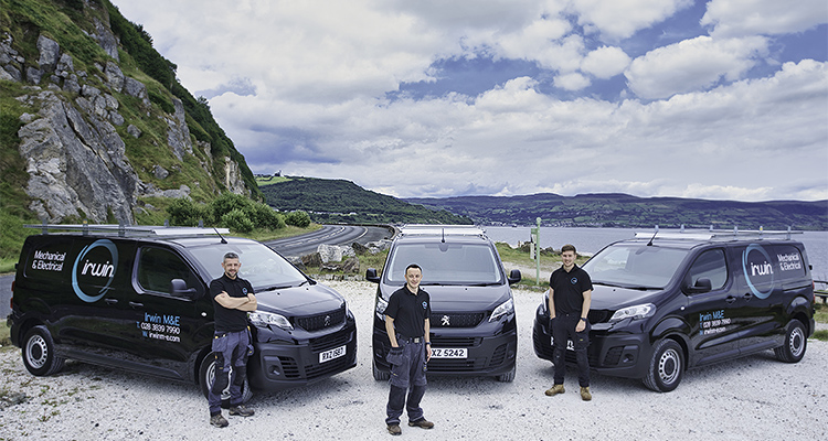 How Irwin M&E has grown from local contractor to leading M&E solutions provider in the UK and Ireland three individuals standing in front of three black vans with the "Irwin M&E" logo, set against a scenic backdrop of mountains and a body of water