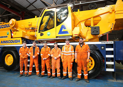 a group of employees from Ainscough Crane Hire, a company that provides lifting solutions in the UK. They are standing in front of a large yellow mobile crane