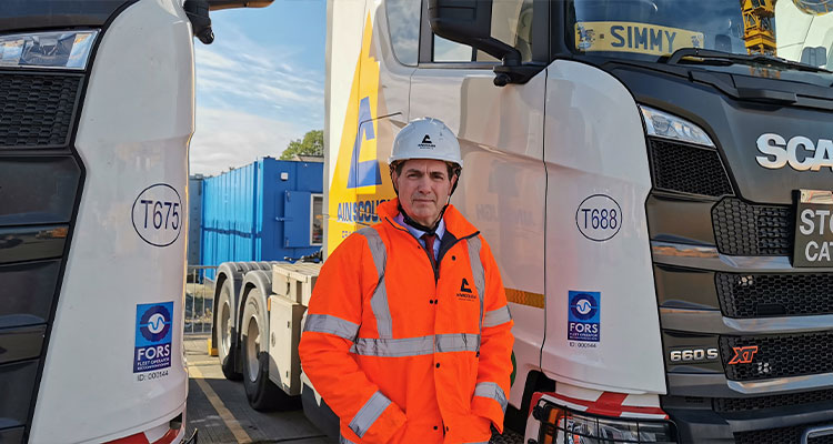 Safety, innovation, and investment have shaped the last 12 months for Ainscough Crane Hire a man in a hard hat and high-visibility jacket standing between two Scania trucks