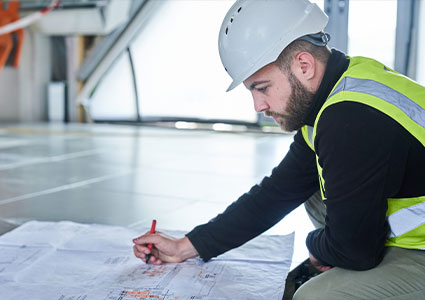 a builder or a civil engineer, wearing a white hard hat and a yellow high-visibility vest over a black long-sleeved shirt. He is kneeling on a polished concrete floor and is focused on a large blueprint or plan spread out on the ground