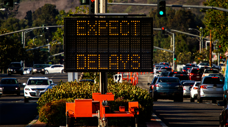 Traffic management sign on a busy road warning of delays during construction works, with vehicles and road infrastructure in an urban setting