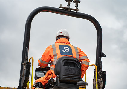 a person in a high-visibility orange jacket and a white hard hat operating heavy machinery, with a blue and white "JB" logo visible on the back of their gear