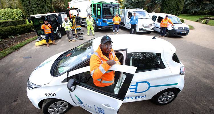a man in an orange high-visibility jacket and a cap, standing next to a white electric car with its door open