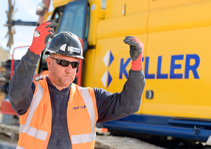 a construction worker giving hand signals in front of a yellow vehicle branded with the Keller logo