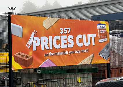 a large, rectangular outdoor advertisement banner mounted on a metal fence structure outside a retail store under a cloudy sky