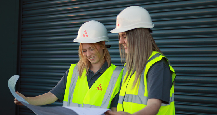 two female construction professionals reviewing building blueprints on-site. They are equipped with essential personal protective equipment (PPE), including custom-branded white hard hats and high-visibility safety vests, posed against a dark gray industrial roller shutter door