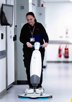 a woman pushing a white and grey i-Mop floor scrubber down a bright, clean hallway