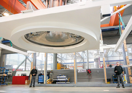 a massive, white, geometric mechanical framework suspended horizontally above the ground. At its core is a large, circular opening containing a reflective, metallic component