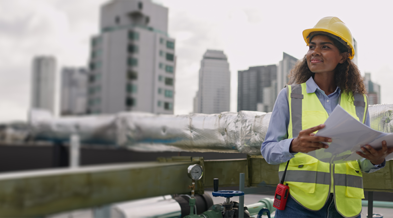 Female construction engineer wearing a yellow hard hat and high-visibility vest reviewing plans on a rooftop construction site, representing UK contractors.