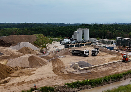 an industrial concrete batching and aggregate supply plant featuring vertical storage silos, stockpiles of sand and gravel, dump trucks, and heavy machinery