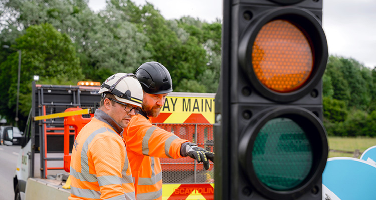 two road maintenance and traffic management workers in high-visibility orange clothing and hard hats setting up or adjusting a temporary traffic light unit on a UK roadway