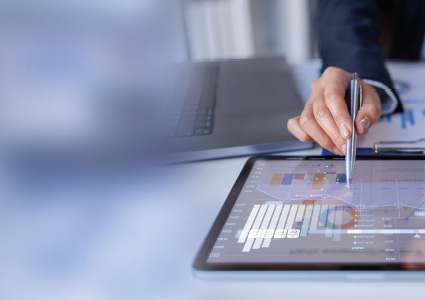 a person using a stylus to review financial data and charts on a tablet device at a desk.