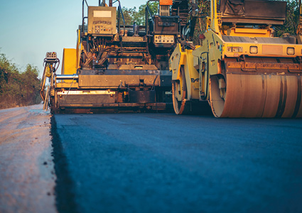 heavy road construction equipment, including an asphalt paver and a smooth-drum road roller, paving a fresh layer of hot-mix asphalt on a highway