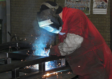 a worker wearing a red protective coverall and a welding helmet while performing metal arc welding on a steel frame