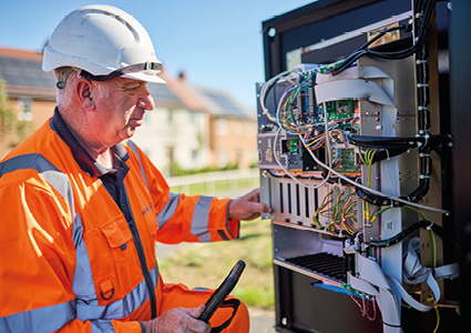 The image shows a telecommunications or utility technician in high-visibility orange workwear and a hard hat inspecting or maintaining an open outdoor broadband/telecom street cabinet