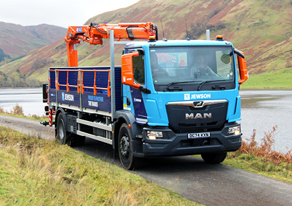 a blue and dark grey MAN flatbed delivery truck driving along a narrow, paved road in a scenic, rural area