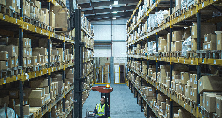 a large industrial warehouse filled with high-rise yellow metal pallet racking systems stacked with cardboard boxes and a worker operating a forklift in the central aisle